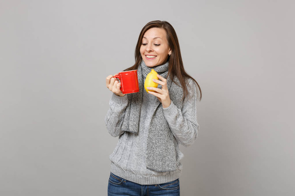 Dość młoda kobieta w szary sweter, szalik trzyma cytryny, czerwony filiżankę herbaty na białym tle na tle szarej ścianie. Moda na zdrowy styl życia, szczere emocje ludzi, koncepcja zimnej pory roku. Makieta do miejsca kopiowania - Zdjęcie, obraz