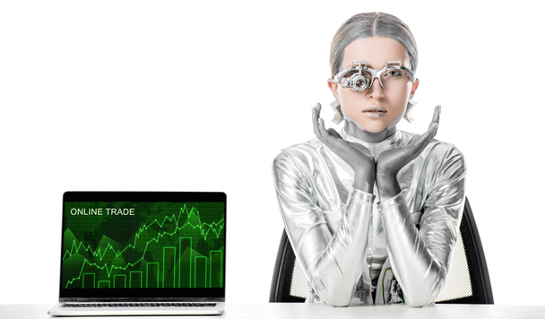 Gümüş robot izole beyaz, geleceğin teknoloji kavramı üzerinde online ticaret cihazı ile dizüstü yakınındaki masada oturan - Fotoğraf, Görsel
