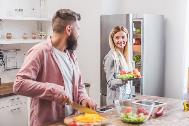 Σύζυγος κοπής λαχανικών και γυναίκα στέκεται κοντά στο ψυγείο - Φωτογραφία, εικόνα