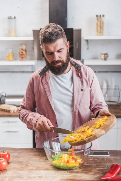 handsome bearded man adding chopped bell pepper to salad at kitchen table - Foto, Bild