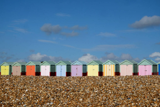 Ряд із десяти барвистих пляж, хатин в черзі у центрі зображення, на передньому плані є гальковий пляж вище знаходиться велика Синє небо з білі хмари світить сонце, Брайтон, Sussex, Сполучені Kingdon, Великобританія - Фото, зображення