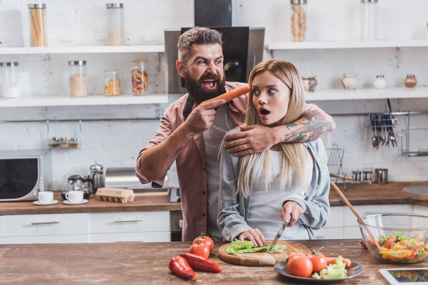 homem engraçado colocando cenoura perto da cabeça da mulher, enquanto a menina assustada cozinhar jantar
 - Foto, Imagem