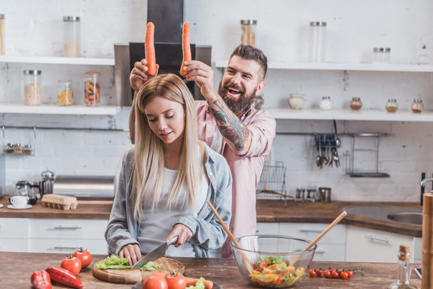 funny man putting carrots on woman head while girl cooking dinner - Zdjęcie, obraz