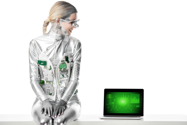 smiling silver robot sitting on table and looking at laptop with business appliance isolated on white, future technology concept - Фото, зображення