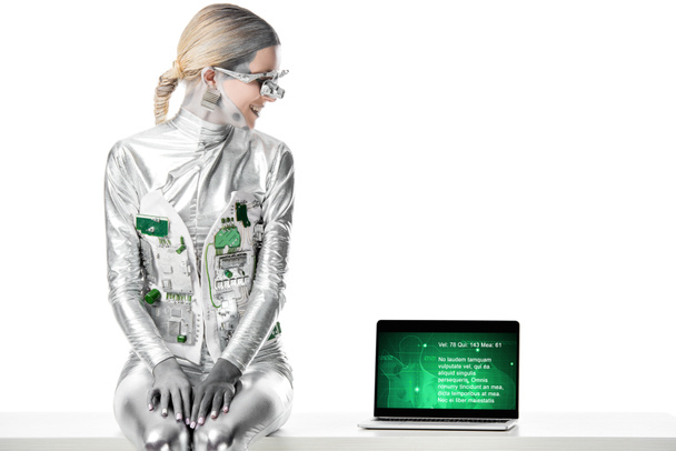 usmívající se stříbrný robot sedí na stole a při pohledu na notebook s lékařský přístroj izolovaných na bílém, budoucí technologický koncept - Fotografie, Obrázek