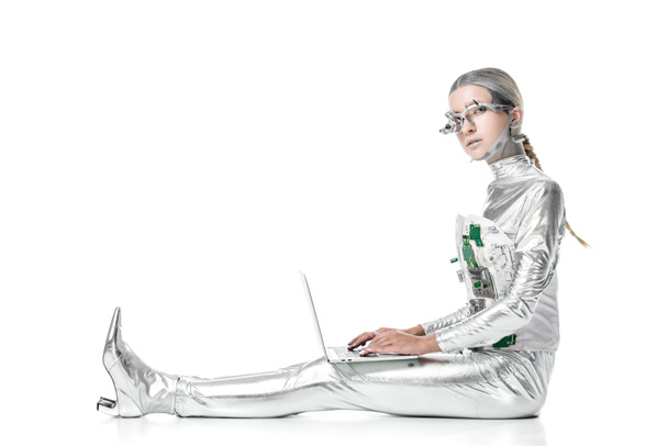 вид сбоку серебряного робота, сидящего с ноутбуком и смотрящего на камеру, изолированную на белом, концепция технологии будущего
 - Фото, изображение