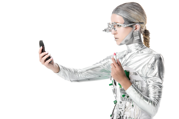 Zijaanzicht van de zilveren robot holding lippenstift en spiegel kijken geïsoleerd op wit, toekomstige technologie concept - Foto, afbeelding