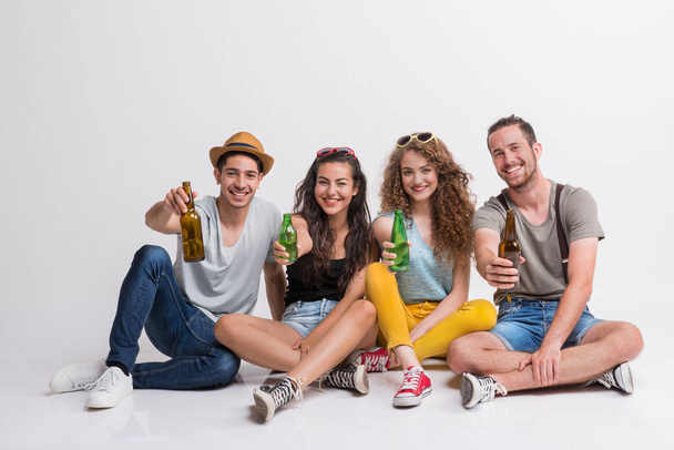 Portrait of joyful young group of friends with bottles sitting on the ground in a studio. - Foto, Bild