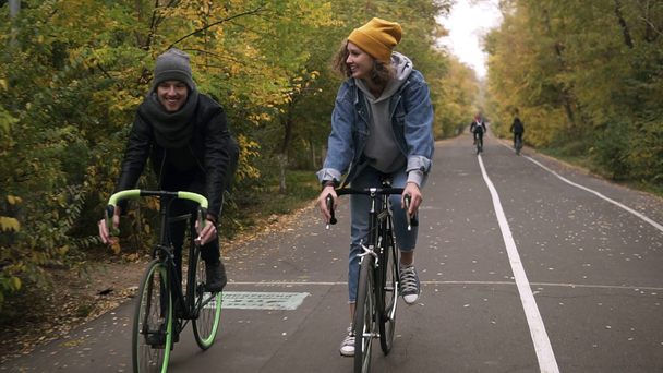Jóvenes amigos sonrientes o parejas jóvenes en sombreros en bicicleta en sus bicicletas de trekking a través del parque de otoño en bicicletas. Hombre y mujer montando bicicletas juntos y hablando. Vista frontal
 - Imágenes, Vídeo
