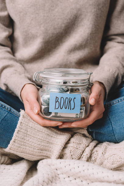 cropped shot of girl holding glass jar with savings and label books - Fotografie, Obrázek