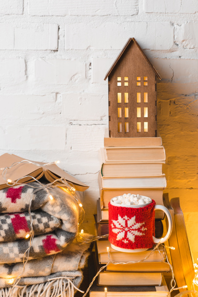 cup of hot chocolate with marshmallows, books and decorative wooden house near white brick wall - Фото, изображение