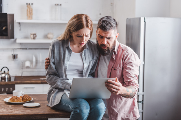 confused young couple looking at laptop screen at kitchen - Photo, Image