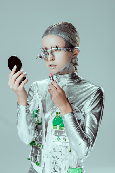 silver robot looking at mirror and holding lipstick isolated on grey, future technology concept - Photo, Image