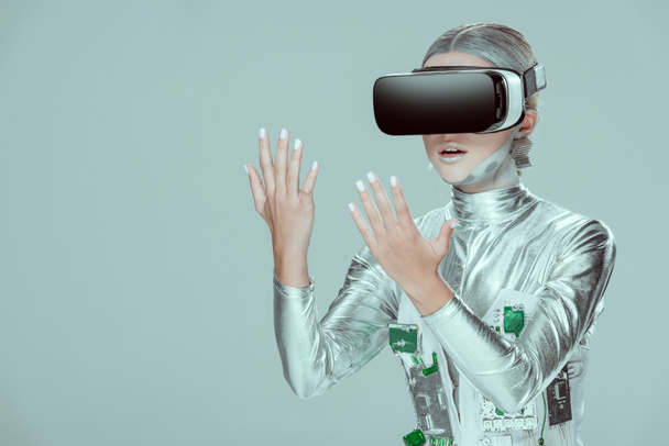 удивленный серебряный робот, смотрящий на руки с гарнитурой виртуальной реальности, изолированной на серой концепции технологии будущего
 - Фото, изображение