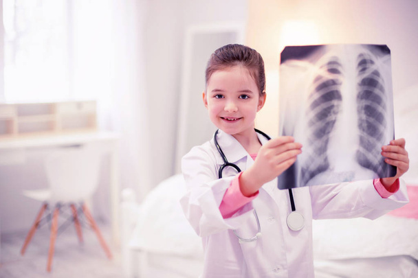 Menina pré-escolar de olhos escuros usando jaqueta branca segurando raio-x dos pulmões
 - Foto, Imagem