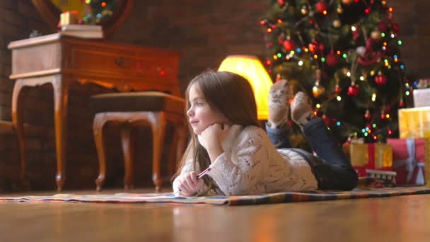 a beautiful little girl in a white knitted sweater lies on the floor, composes and writes a letter to Santa Claus, in the background a festive Christmas tree and a host of gifts - Materiaali, video
