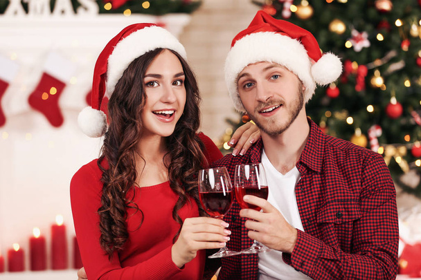 Jovem casal feliz com vinho em copos sentado em casa perto da árvore de Natal
 - Foto, Imagem