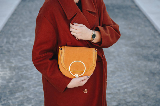 Streetstyle mode details. Close-up, jonge mode blogger dragen bruine winterjas, en een zwarte en gouden analoge pols horloge. de tijd, een mooie suède lederen tas houden controleren. - Foto, afbeelding