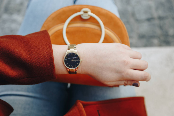 Modedetails im Streetstyle. Nahaufnahme, junge Modebloggerin in braunem Wintermantel und schwarz-goldener analoger Armbanduhr. Zeitkontrolle, in der Hand eine schöne Wildledertasche. - Foto, Bild