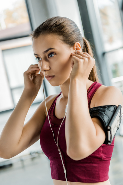 portrait of fit sportswoman in smartphone armband putting on earphones at gym - Foto, Imagen