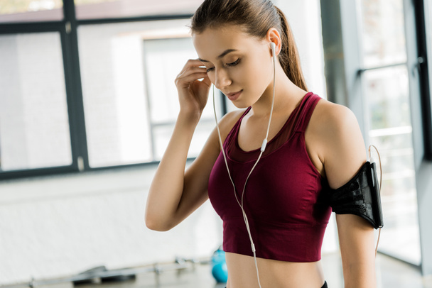 schöne schlanke Sportlerin in Smartphone-Armbinde, die im Fitnessstudio Kopfhörer aufsetzt - Foto, Bild