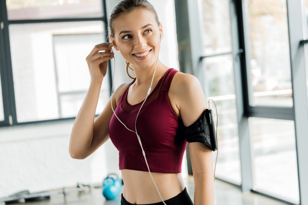 beautiful smiling sportswoman in smartphone armband putting on earphones at gym - Foto, Imagem