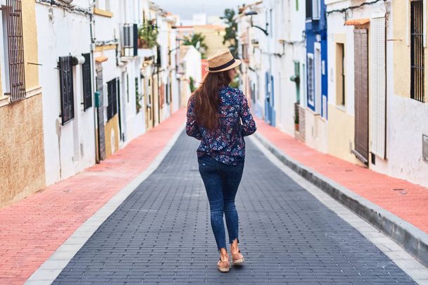Back view of a young long-haired woman in hat walking down the street with small colorful buildings. - Fotoğraf, Görsel