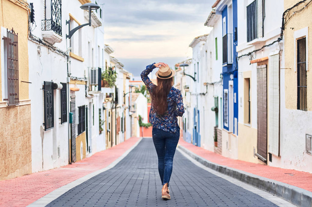 Back view of a young long-haired woman in hat walking down the street with small colorful buildings. - Zdjęcie, obraz