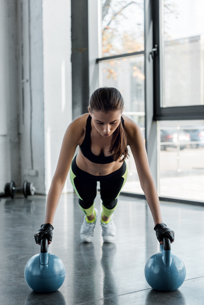 focused sportswoman in weightlifting gloves doing plank exercise on kettlebells at sports center - Φωτογραφία, εικόνα