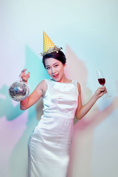 Porträt einer glücklichen schönen Frau im weißen Kleid, die Champagner trinkt, während sie mit Discokugel auf hellem Hintergrund steht - Foto, Bild