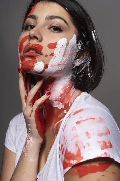Beautiful brunette girl wearing a white t-shirt stained with paint touches her face and neck with her hand. Handprint of the palm and strokes of paint on her face. Conceptual, fashion design - Photo, Image