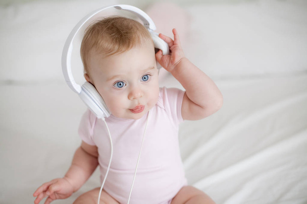 Sinisilmäinen tyttö vaaleanpunaisessa puvussa kuulokkeet sängyllä. Tyttö leikkii kuulokkeilla. Kodikas. Kotiin
. - Valokuva, kuva