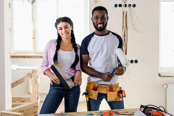 šťastný mladý pár s nástroji při provádění rekonstrukce domova při pohledu na fotoaparát - Fotografie, Obrázek