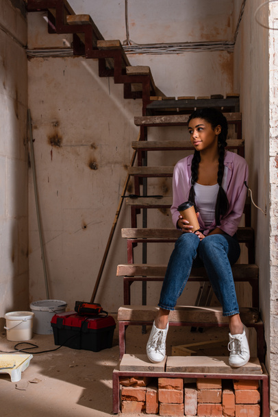 όμορφη νεαρή γυναίκα που κάθεται σε σκαλοπάτια με χαρτί φλιτζάνι καφέ κατά τη διάρκεια της ανακαίνισης του σπιτιού - Φωτογραφία, εικόνα