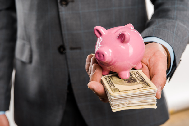 close-up partial view of businessman holding dollar banknotes and piggy bank - Foto, imagen