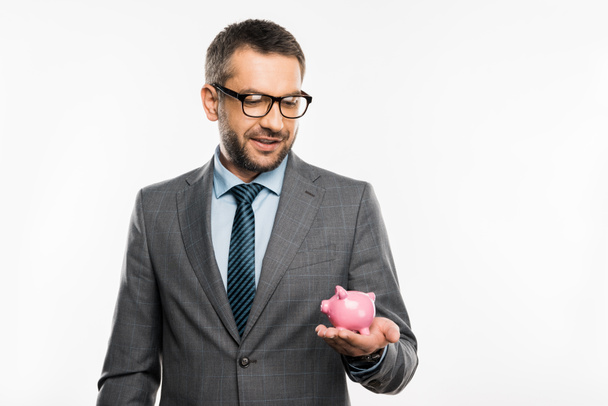 handsome businessman in suit and eyeglasses holding piggy bank isolated on white - Фото, изображение