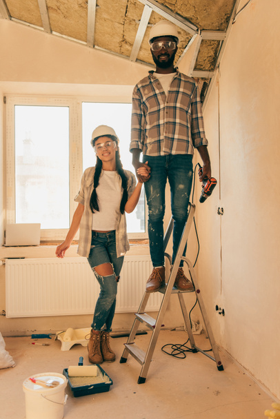 homem americano africano em chapéu duro e óculos de pé com broca de energia na escada, enquanto sua namorada de pé perto durante a renovação de casa
 - Foto, Imagem