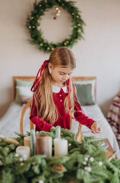 girl in a burgundy dress decorates a Christmas tree in a rustic room - Foto, immagini