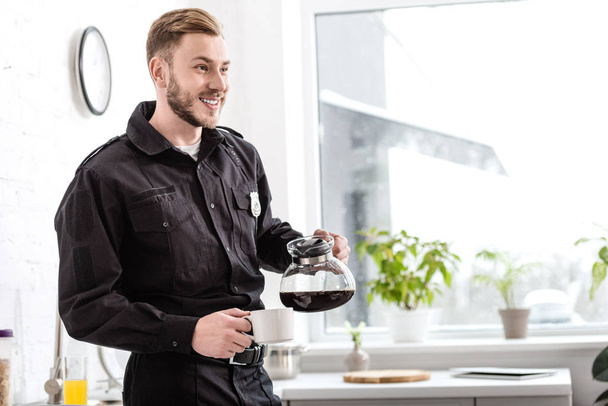 hombre sonriente en uniforme de policía vertiendo café en la taza de la tetera cerca de la ventana de la cocina
 - Foto, Imagen