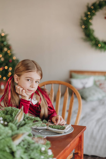 girl in a burgundy dress decorates a Christmas tree in a rustic room - Фото, изображение