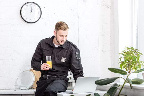 Молодой полицейский сидит за столом со стаканом апельсинового сока и пользуется ноутбуком на кухне
 - Фото, изображение