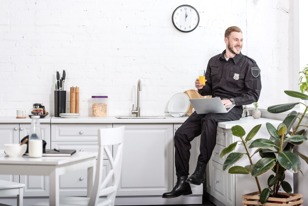 улыбающийся мужчина в полицейской форме сидит на кухонном столе, пьет апельсиновый сок и пользуется ноутбуком
 - Фото, изображение