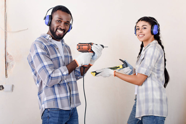 laughing young african american couple in protective headphones posing with drill and hammer during renovation of new home - Photo, Image