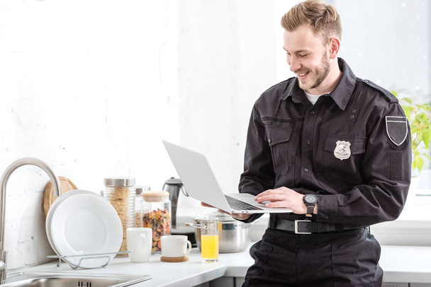 smiling police officer standing and using laptop at kitchen - Photo, Image