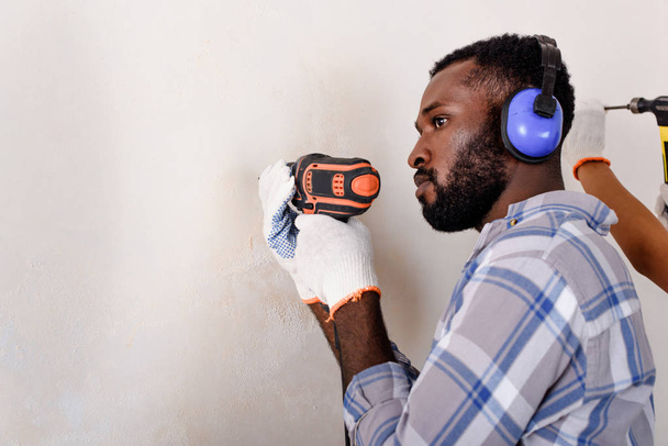 vista lateral del hombre afroamericano reflexivo en auriculares protectores que trabajan con taladro eléctrico en casa
 - Foto, imagen