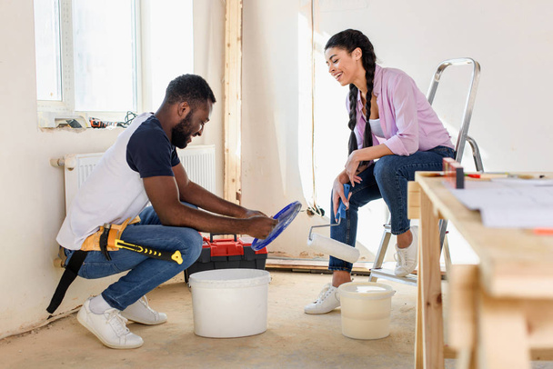 happy young man sitting near bucket with paint while his girlfriend sitting with paint roller during renovation at home - Fotoğraf, Görsel