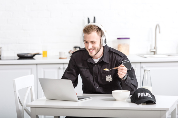 Красивый полицейский ест кукурузные хлопья и пользуется ноутбуком на кухне
 - Фото, изображение