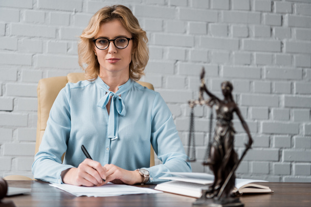 confident female lawyer in eyeglasses sitting at workplace and looking at camera - Photo, Image
