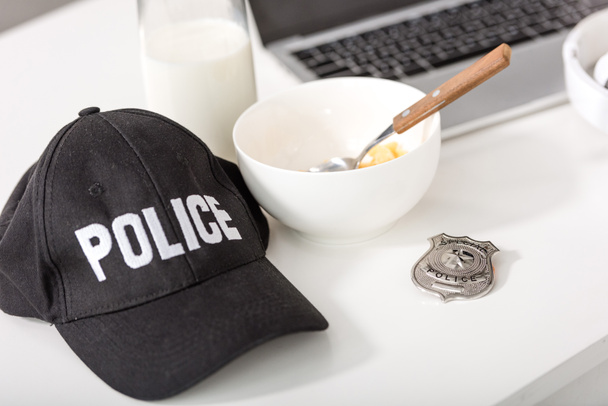 police cap, bowl with cornflakes, police badge and laptop on table - Фото, изображение