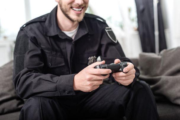 oříznutý pohled usmívající se policista s gamepad sedí na gauči - Fotografie, Obrázek